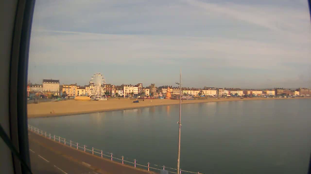 A calm body of water stretches before a sandy beach lined with buildings, a Ferris wheel, and other fairground attractions under a mostly clear sky.