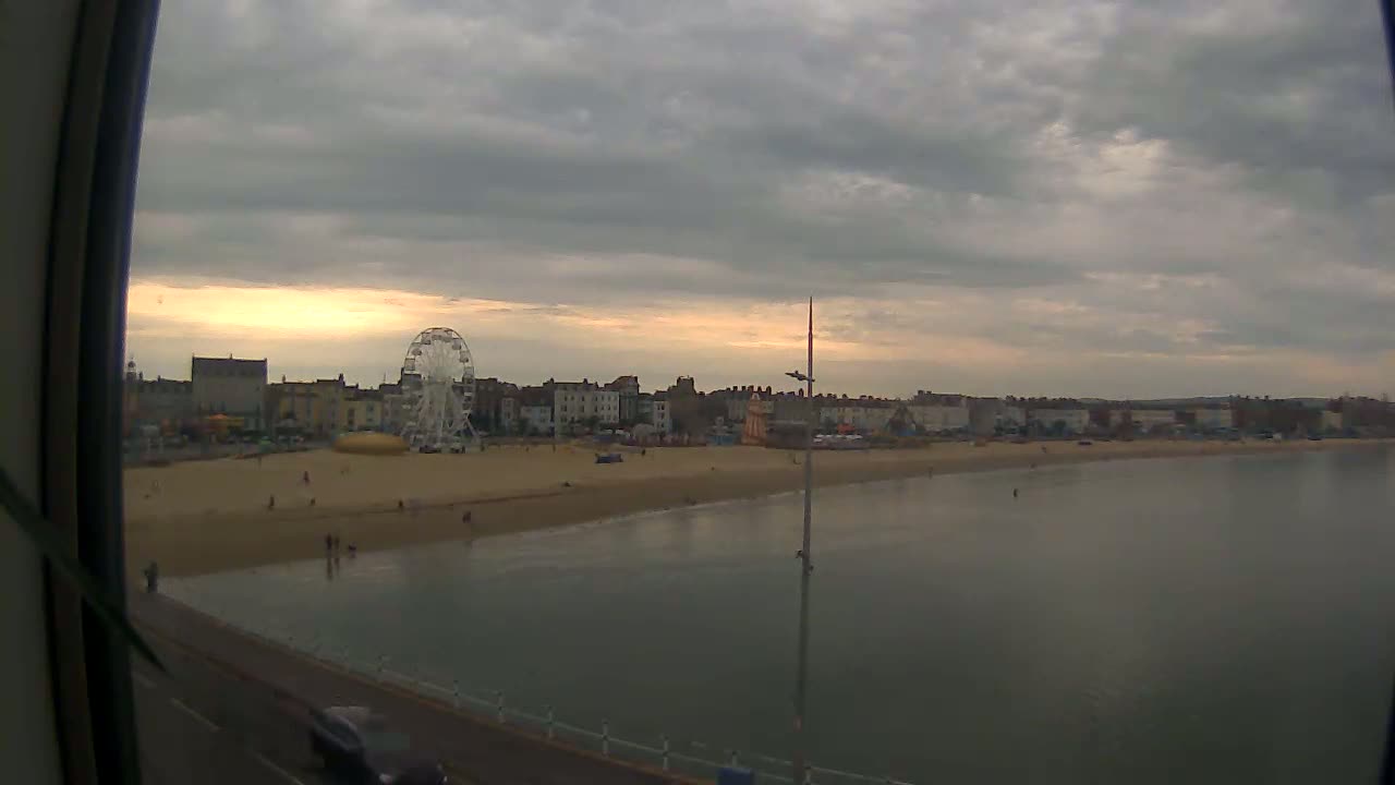A Ferris wheel sits on a beach next to buildings under an overcast sky, with calm water in the foreground.