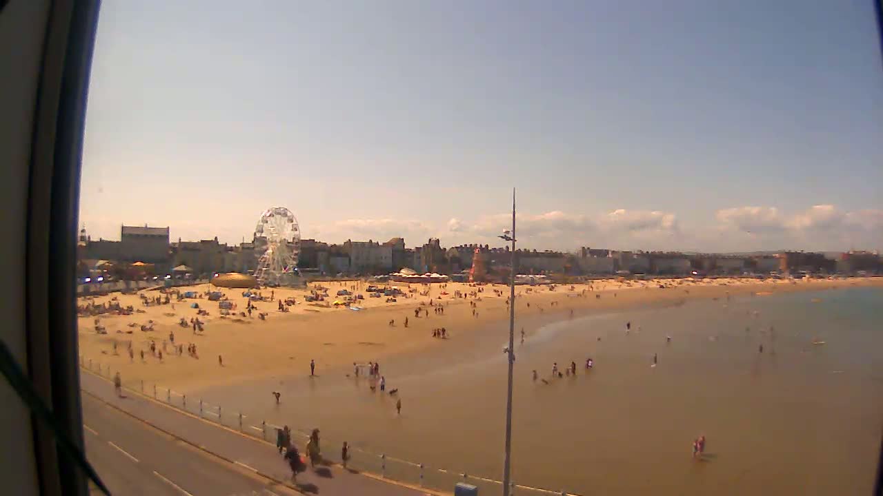 A sandy beach with many people on it and in the shallow water, a Ferris wheel, and buildings in the background on a sunny day with a few clouds.