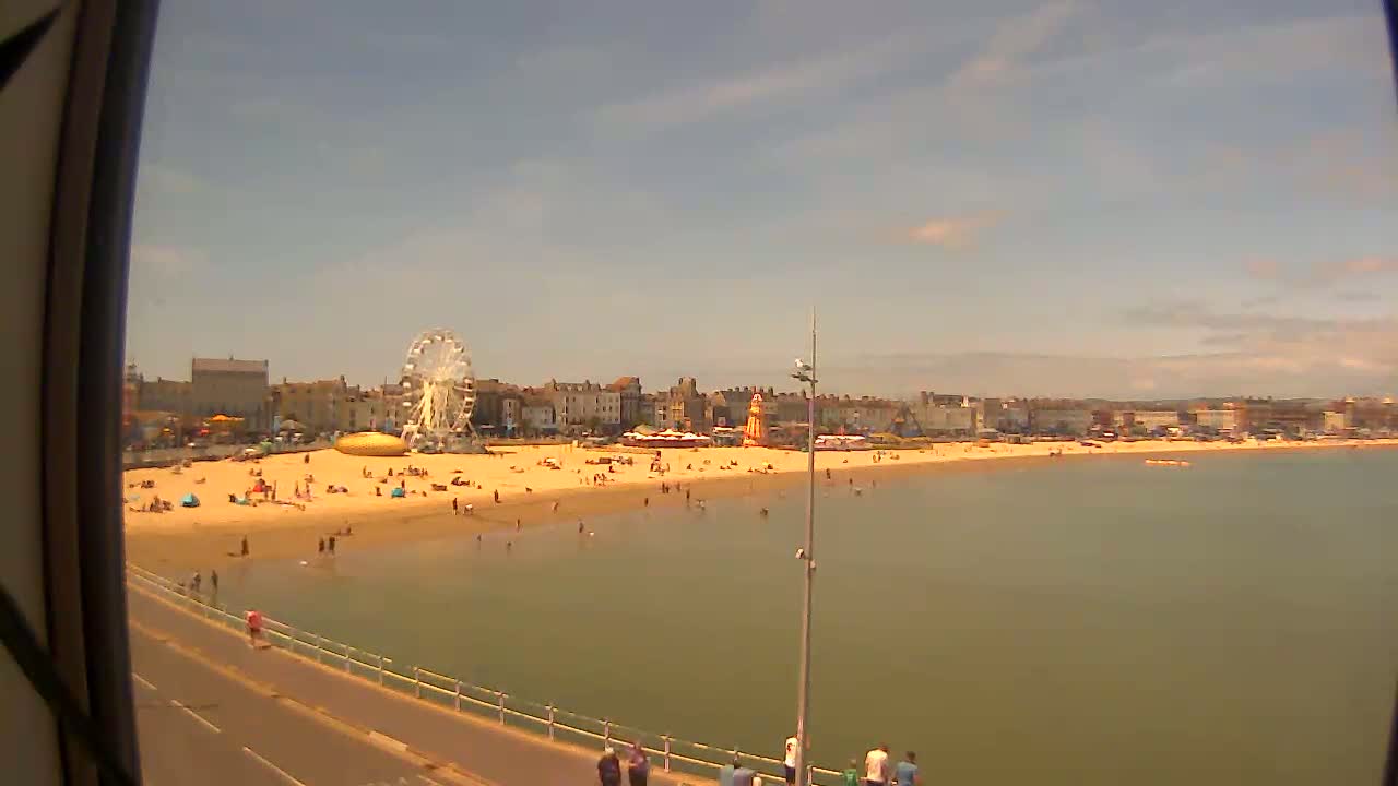 A sunny day at a beach shows a Ferris wheel and other attractions near a sandy shore with many people, and calm, shallow water extending to a distant town.