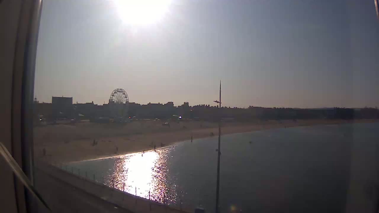 A sunny day shows a beach with calm water, a Ferris wheel, and buildings in the background.