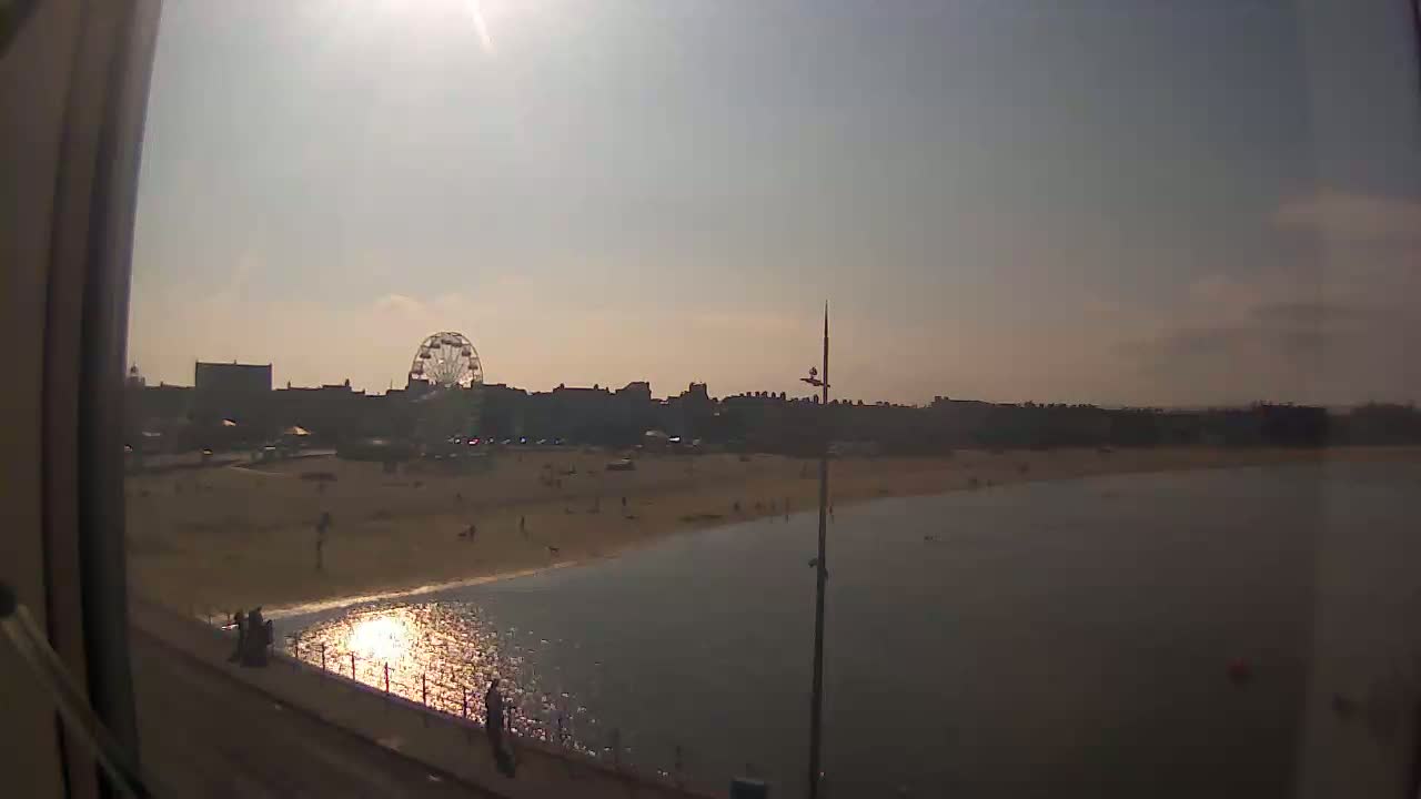 A sunny day shows a beach with a Ferris wheel in the background, calm water, and a few people scattered on the sand.