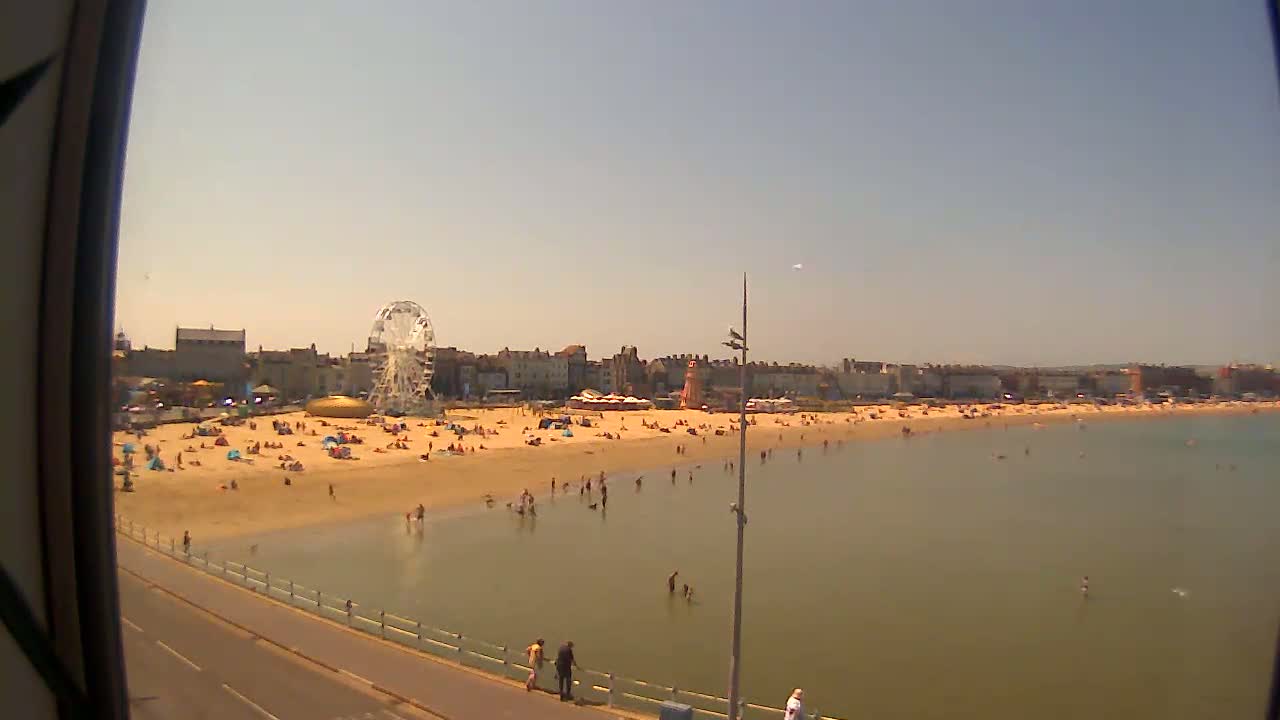 A sunny day at a beach shows a sandy shore with many people, shallow calm water, a Ferris wheel, and buildings in the background.