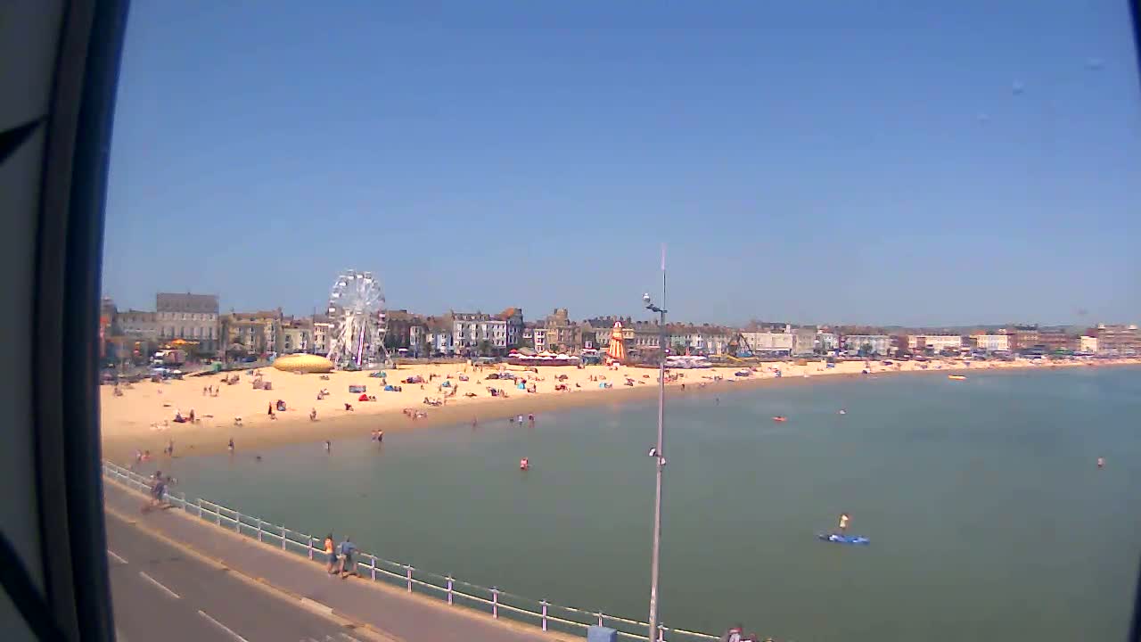 A sandy beach with many people on it, a Ferris wheel, and buildings behind it is shown on a sunny day.