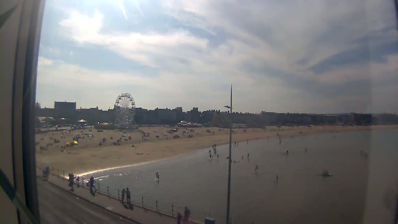 A mostly sunny day at a beach with a Ferris wheel in the background, many people are on the beach and in the shallow water.