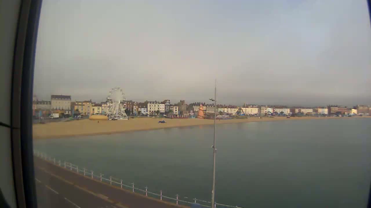 A sandy beach with buildings and a Ferris wheel is visible across calm water under an overcast sky.