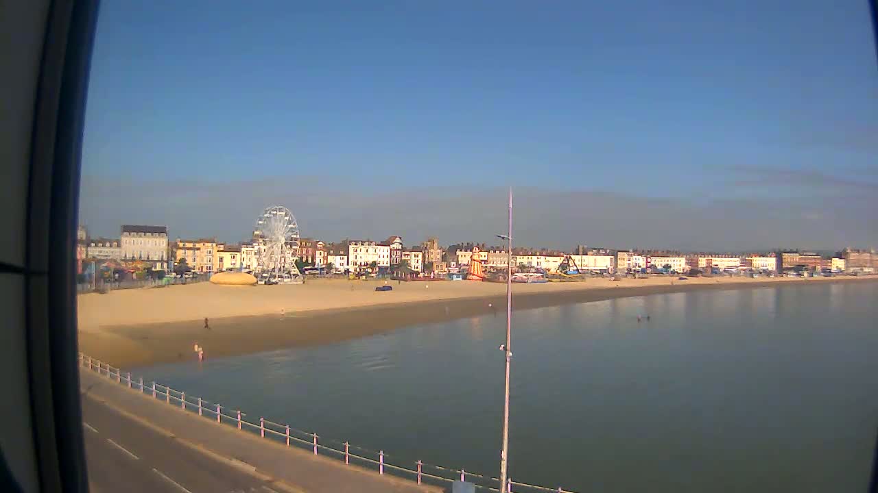 A sandy beach with a Ferris wheel and several buildings stretches along a calm body of water under a clear blue sky.