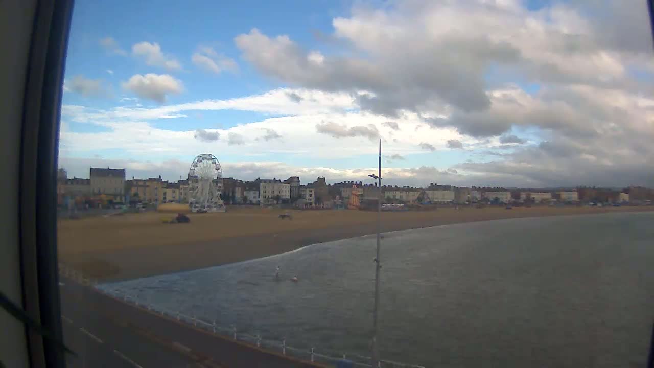 A mostly cloudy day reveals a beach with a Ferris wheel and seaside buildings, next to a calm body of water.