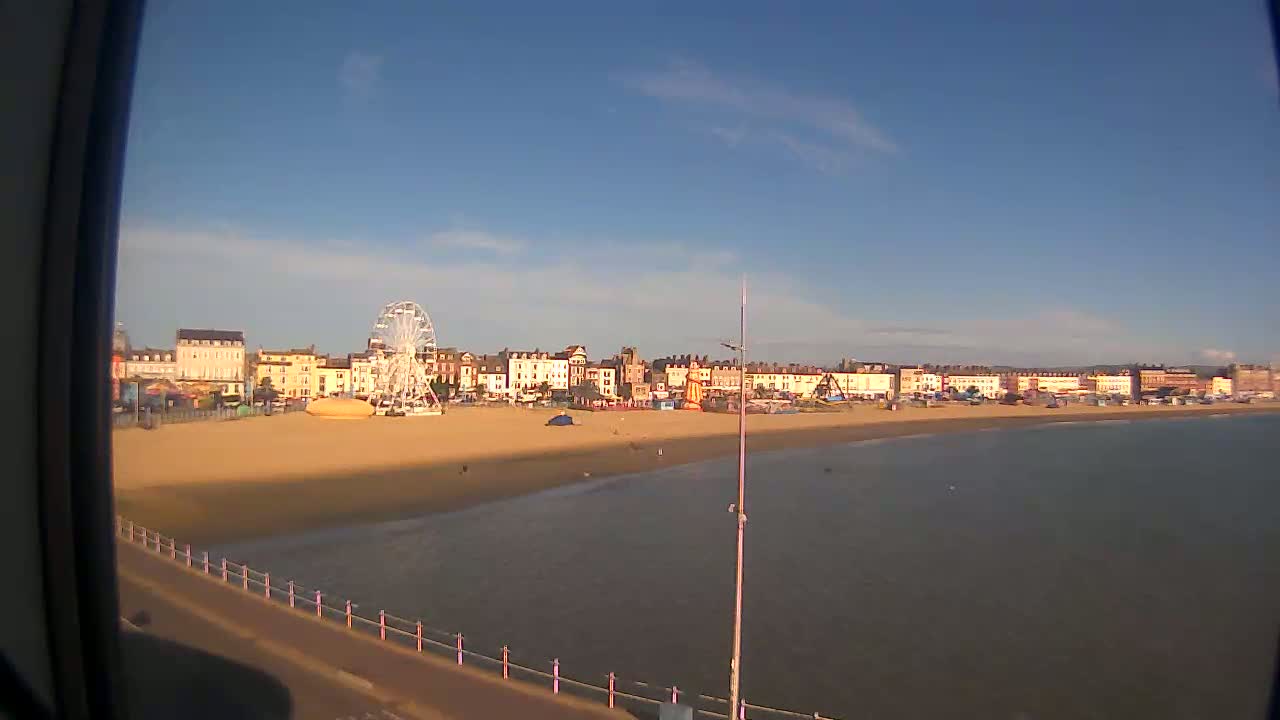 A Ferris wheel sits on a sandy beach next to a row of buildings overlooking calm water under a clear, sunny sky.