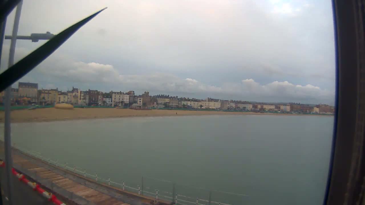 A wide coastal view shows a sandy beach bordered by a line of multi-story buildings and calm grey water, all under an overcast and cloudy sky, with parts of a pier visible in the foreground.