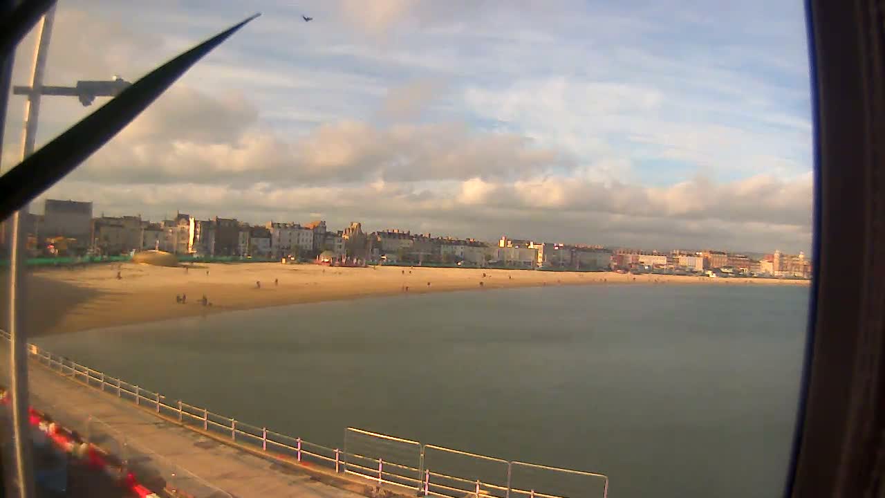 A wide coastal view shows a sandy beach bordered by a line of multi-story buildings and calm grey water, all under an overcast and cloudy sky, with parts of a pier visible in the foreground.