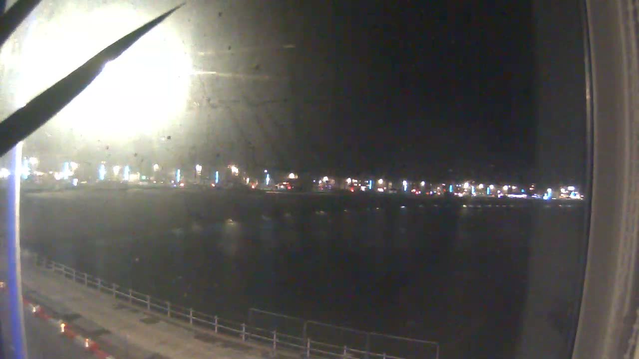 A wide coastal view shows a sandy beach bordered by a line of multi-story buildings and calm grey water, all under an overcast and cloudy sky, with parts of a pier visible in the foreground.