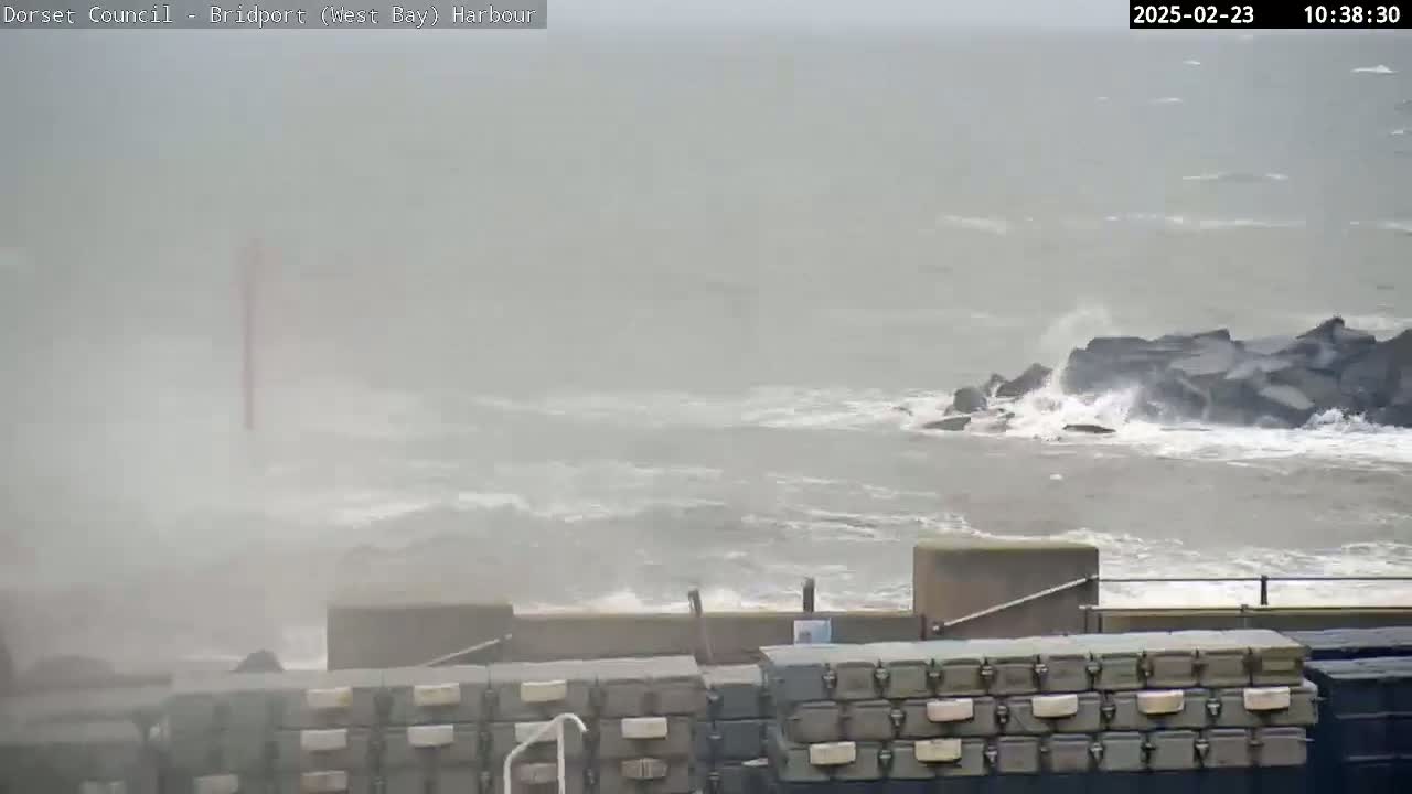 A hazy, overcast day shows rough seas crashing against a rocky breakwater, with several stacks of storage containers visible in the foreground.