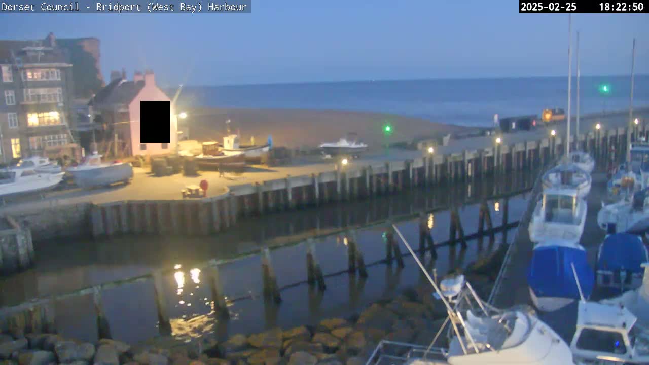 A harbor at twilight shows several boats moored at a dock, with a calm sea and a sandy beach in the background.