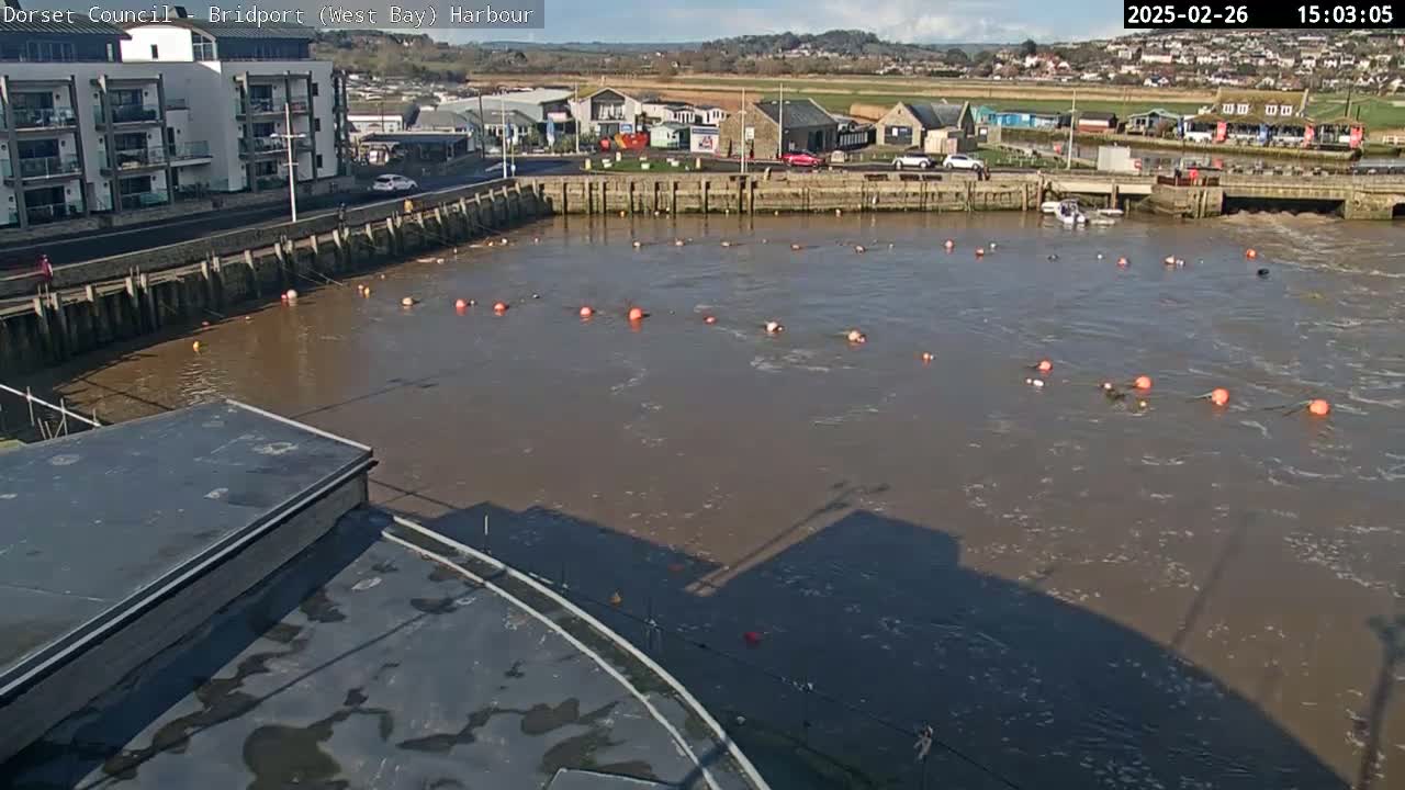 A harbor with muddy brown water containing several orange buoys is seen from an elevated vantage point on a partly sunny day.