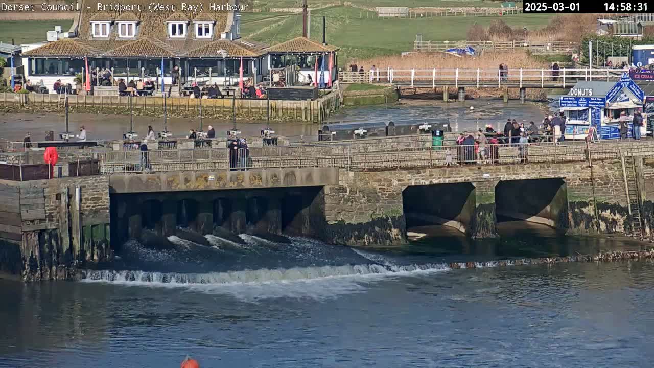 Water flows from a stone structure over a low dam into a harbor on a sunny day, with people gathered near a building and food stand.