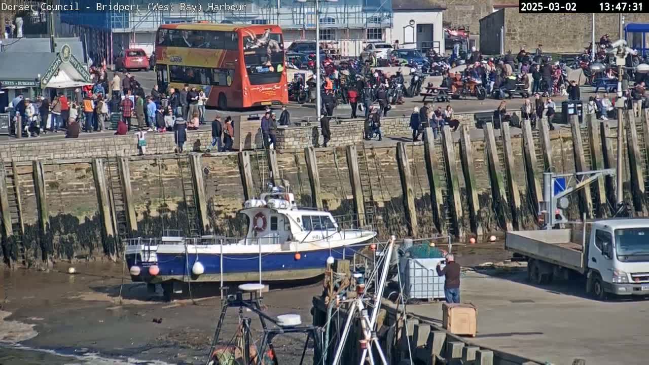 A harbor scene on a sunny day shows a small fishing boat resting on the mud, a double-decker bus parked nearby, many people gathered along a stone wall, and a flatbed truck parked on the pavement.