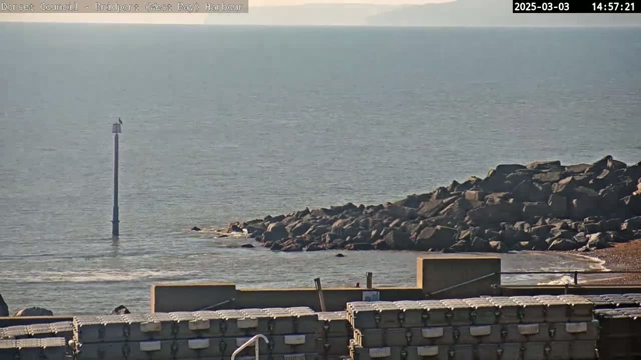 A calm sea with a rocky outcrop and a tall, slender pole in the water, under a mostly clear sky.