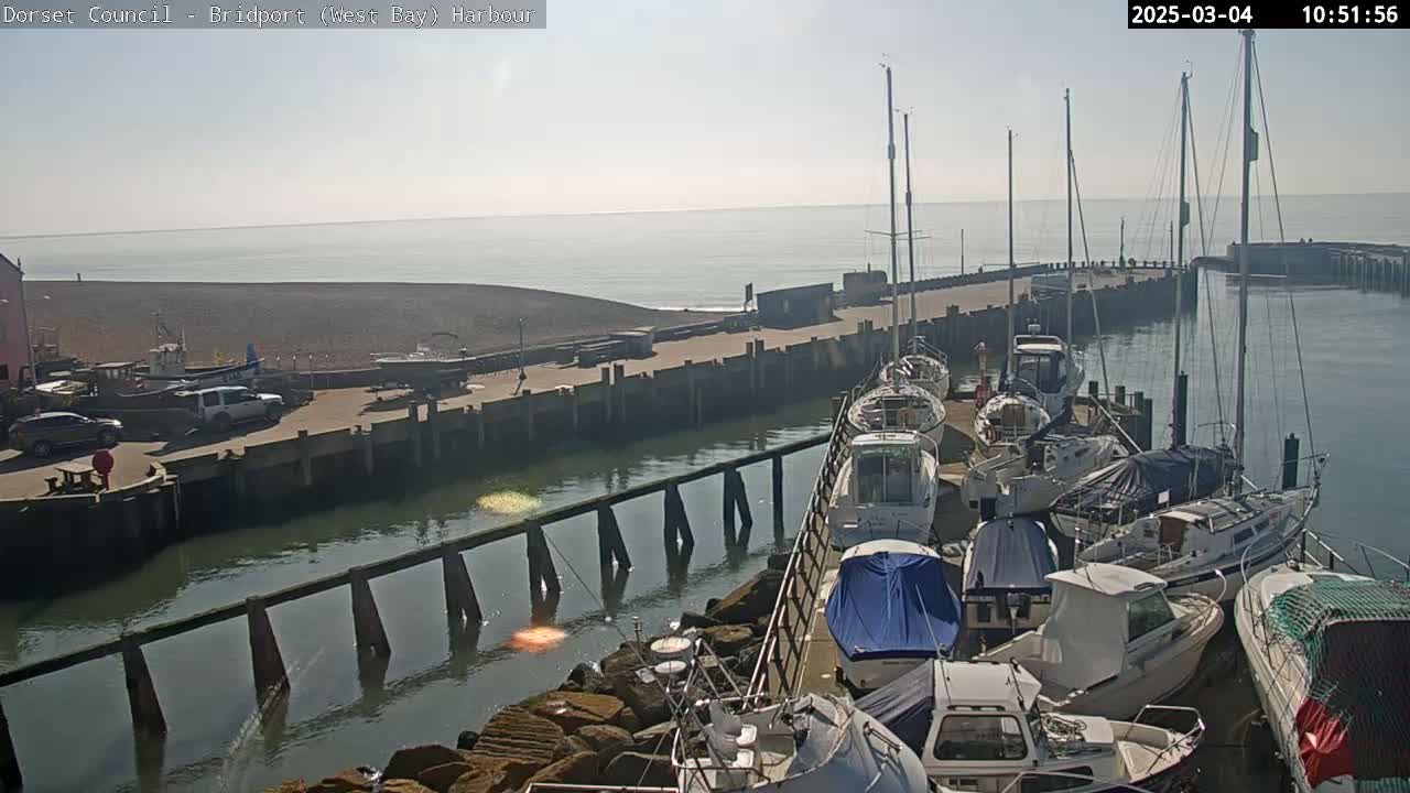 A harbor filled with numerous boats on a sunny day, with a pebble beach and calm water visible in the background.
