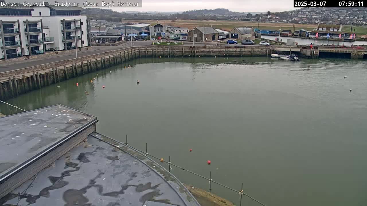 A harbor with calm, gray water is seen from above, featuring a building with a flat roof and a waterfront area with some cars parked nearby under an overcast sky.