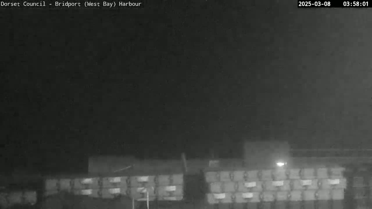 A nighttime, low-light view shows a large stack of rectangular objects, possibly lumber, under a dark sky.