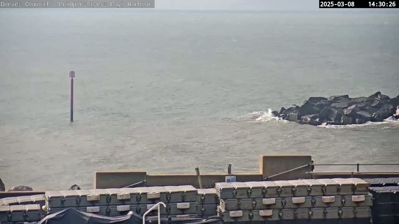 A hazy, somewhat choppy body of water features a navigational marker and a rocky outcropping with small waves crashing against it, with stacked containers visible in the foreground.