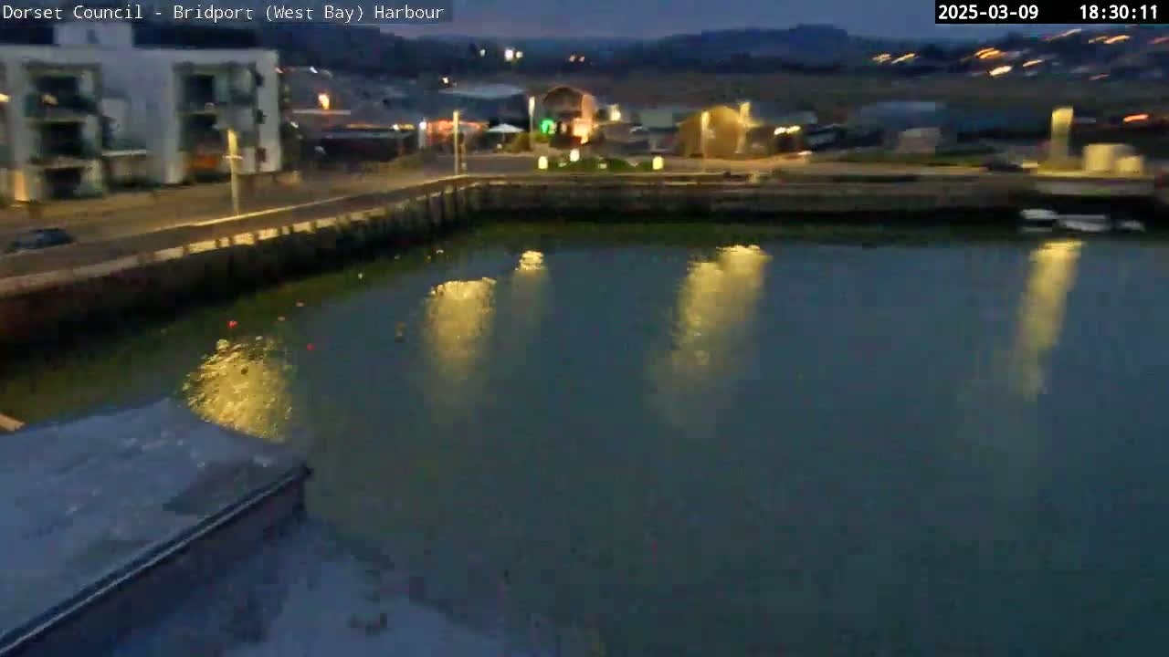 A calm harbor at dusk reflects the lights of buildings and streetlights on its dark waters.