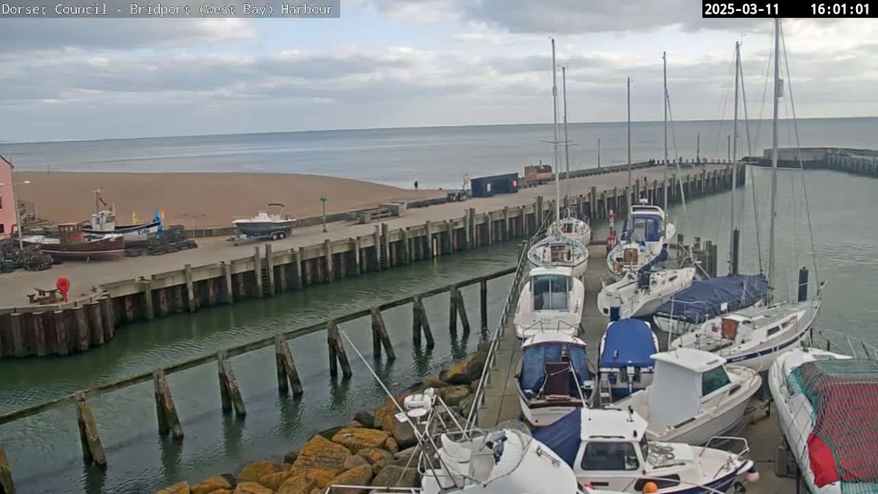 A harbor filled with sailboats and motorboats under a partly cloudy sky, next to a sandy beach and calm sea.