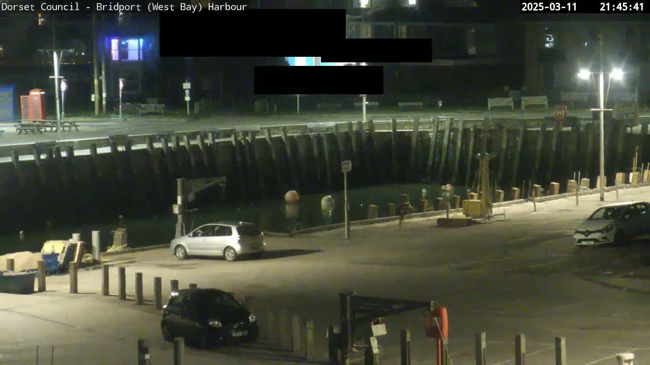 A nighttime harbor scene shows a few cars parked in an empty lot next to a dark water basin, bordered by a wooden wall under bright streetlights.
