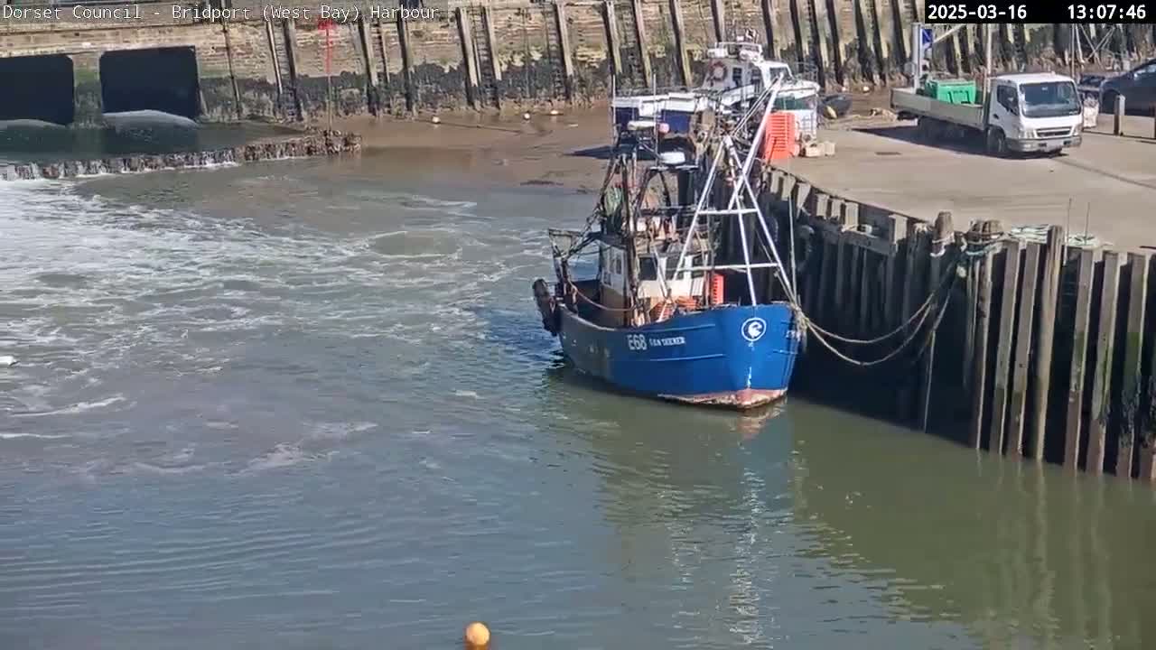 A blue fishing boat is docked at a harbor next to a stone wall on a sunny day with choppy water.