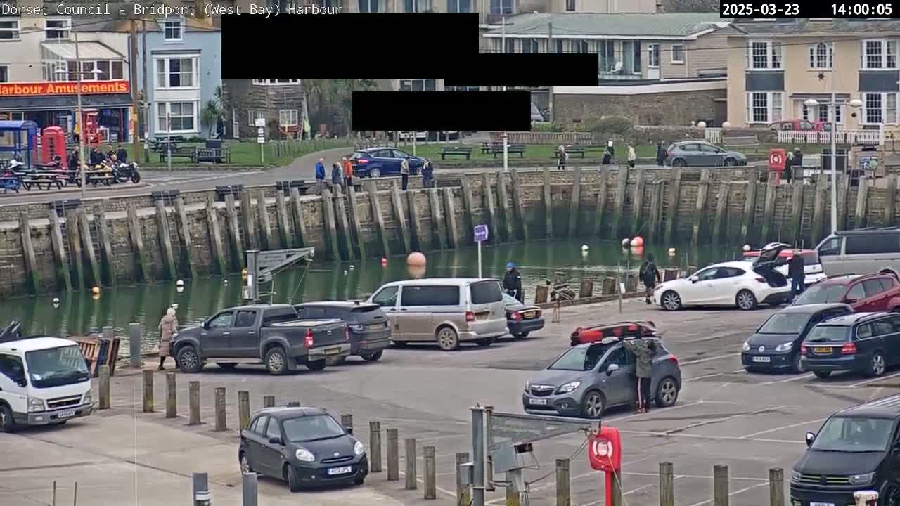 A harbor scene shows a parking lot filled with cars next to a calm body of water and a stone wall, with people walking nearby and buildings in the background on a cloudy day.