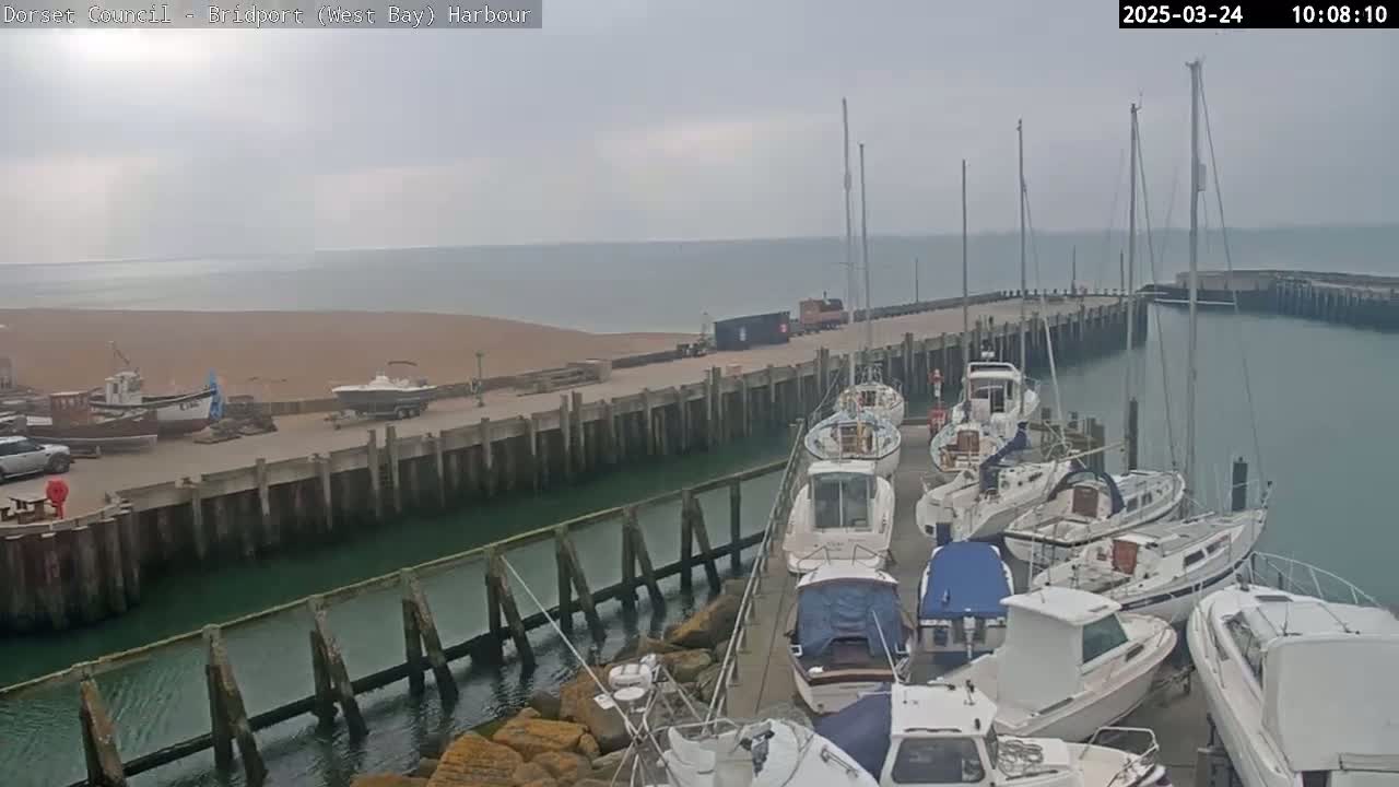 A harbor filled with docked boats and yachts on an overcast day, with a sandy beach visible in the background.