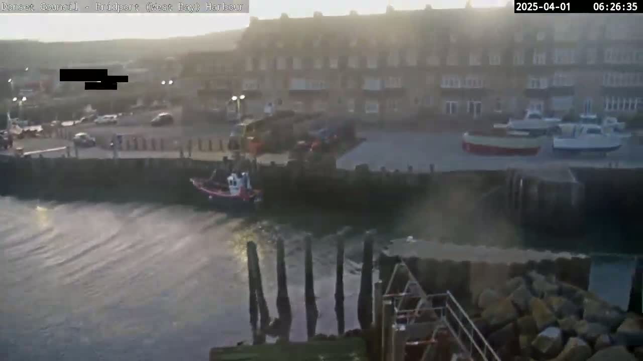 A harbor scene at sunrise shows several boats moored near a stone building, with calm water lapping against a stone seawall.