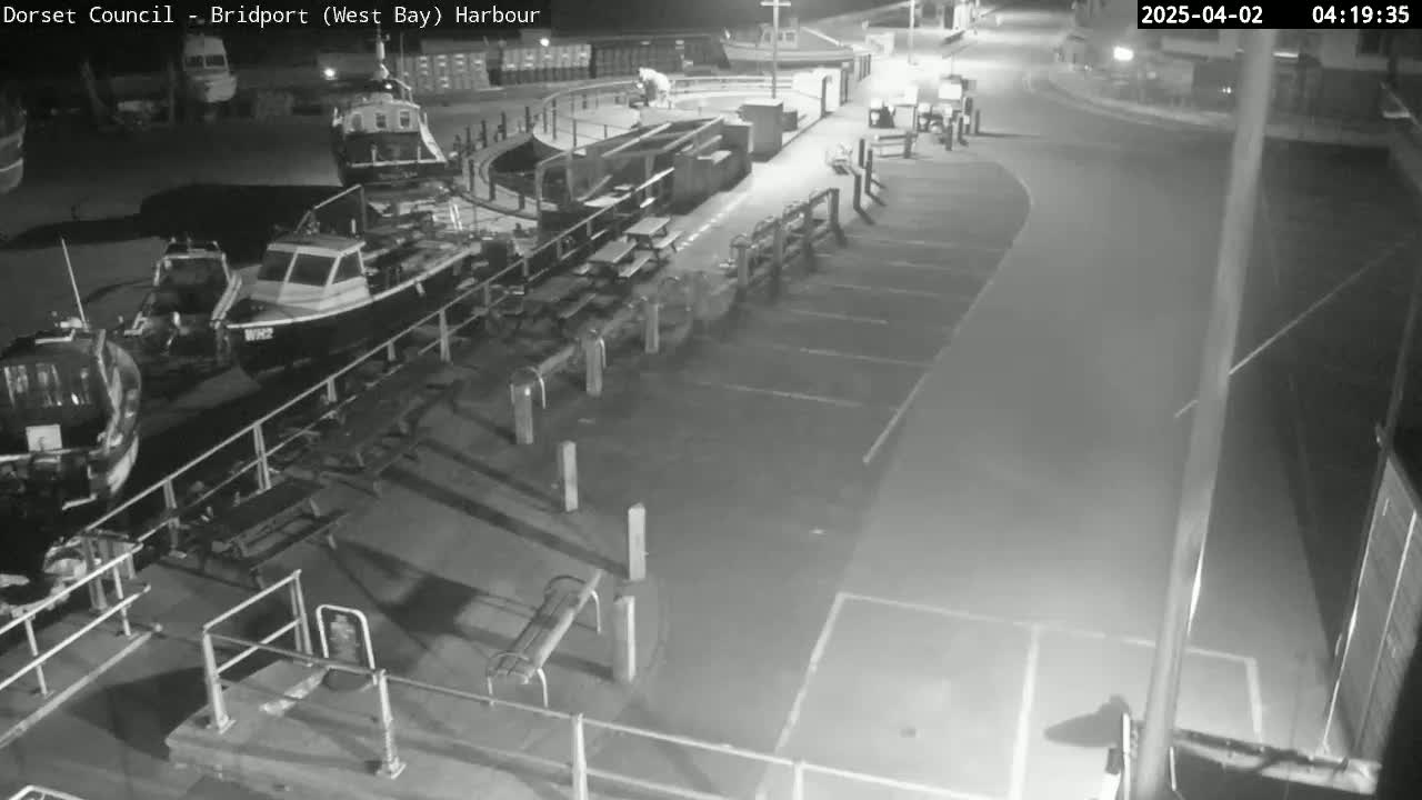 At night, several small boats are moored at a harbor next to an empty parking lot with picnic tables.