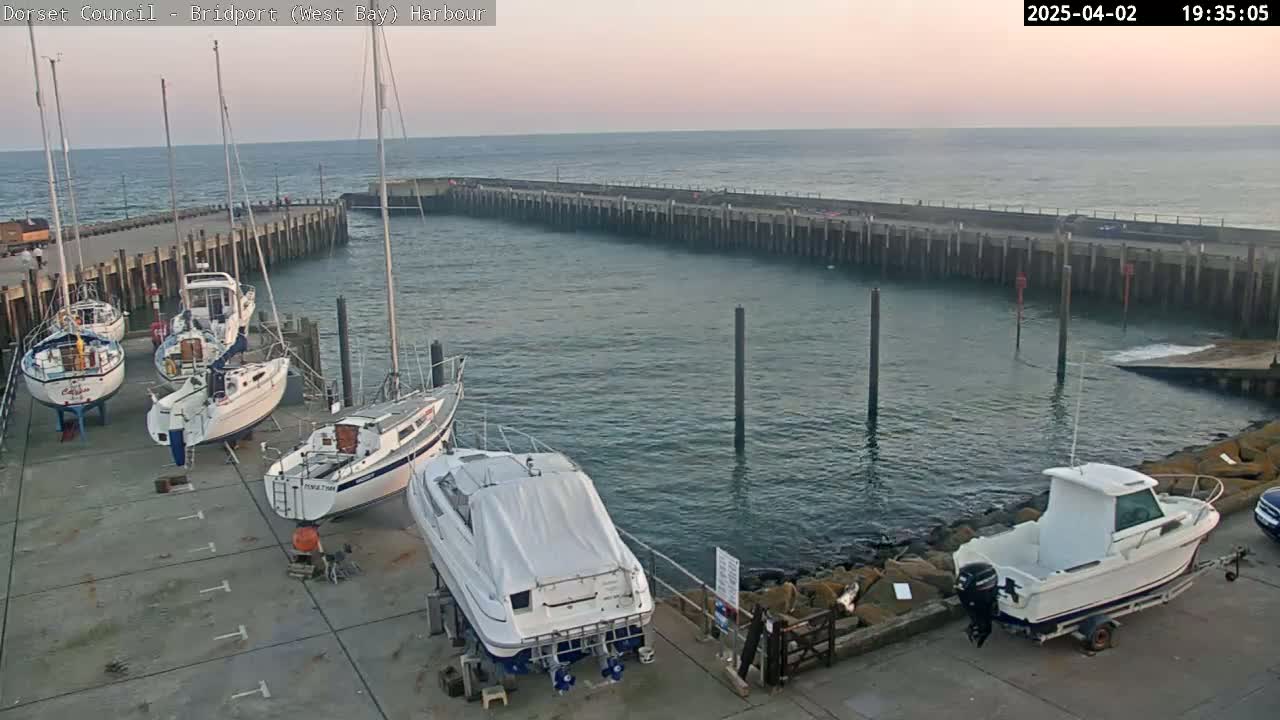 Several sailboats and motorboats are docked at a harbor, with calm water and a twilight sky in the background.