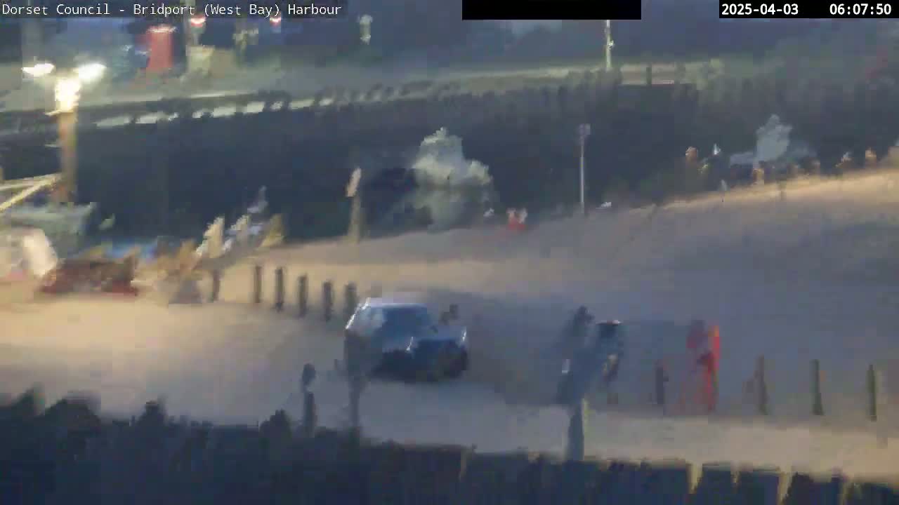A dark-colored car drives on a sandy area near a harbor wall under hazy, bright conditions.