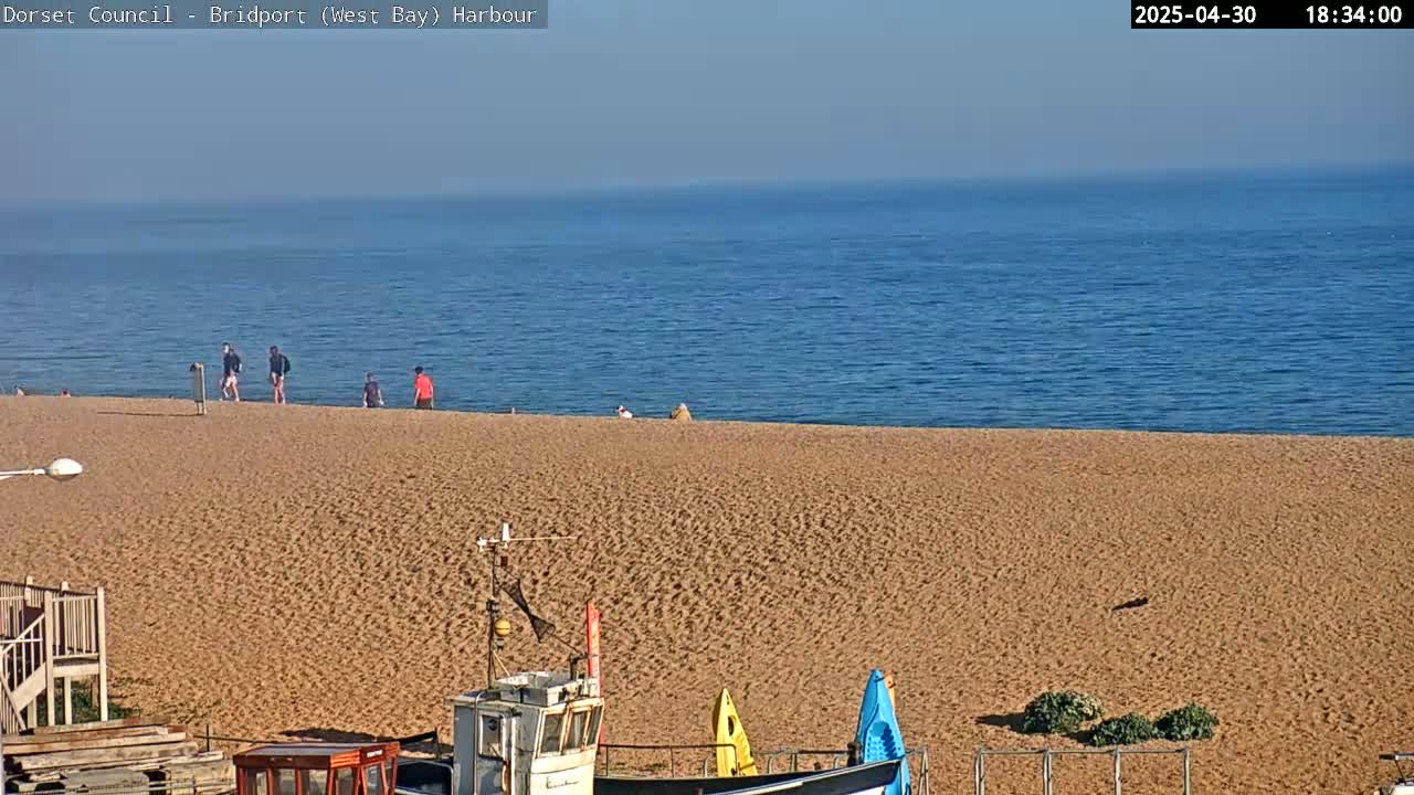 A sandy beach stretches to a calm blue sea under a clear sky, with a few people walking on the shore and small boats visible in the foreground.
