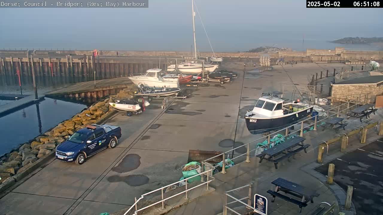 Several boats are docked at a harbor on a hazy morning, with a pickup truck parked nearby and picnic tables visible on the quay.