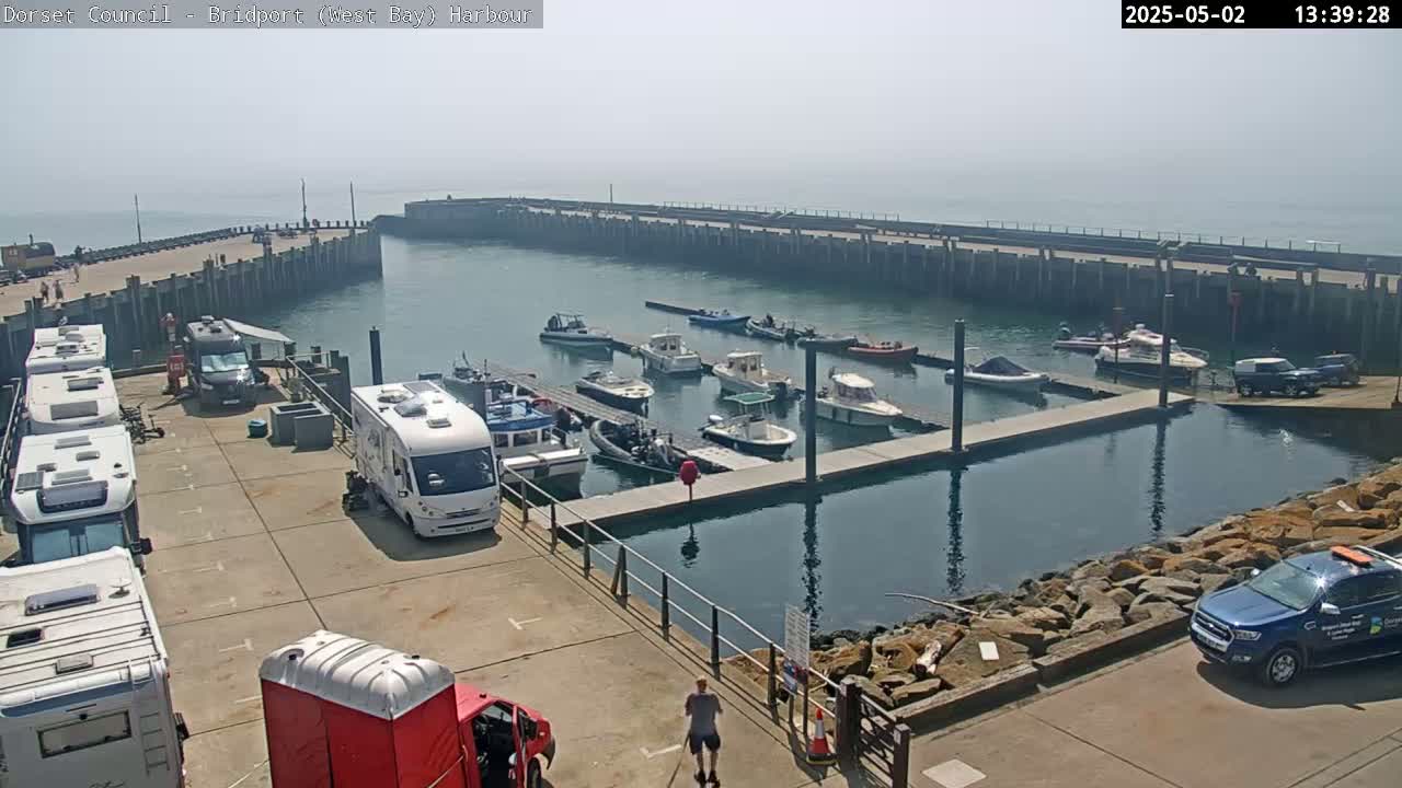 A harbor with numerous boats docked, several campervans parked on the quay, and a hazy, sunny day.