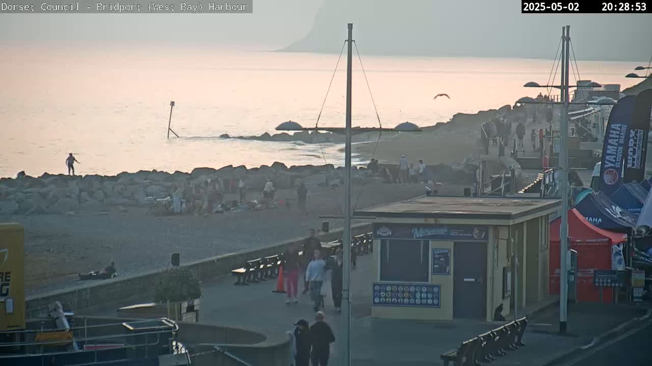 A pebble beach with a few people walking along a promenade, near a small building, under a soft sunset sky.