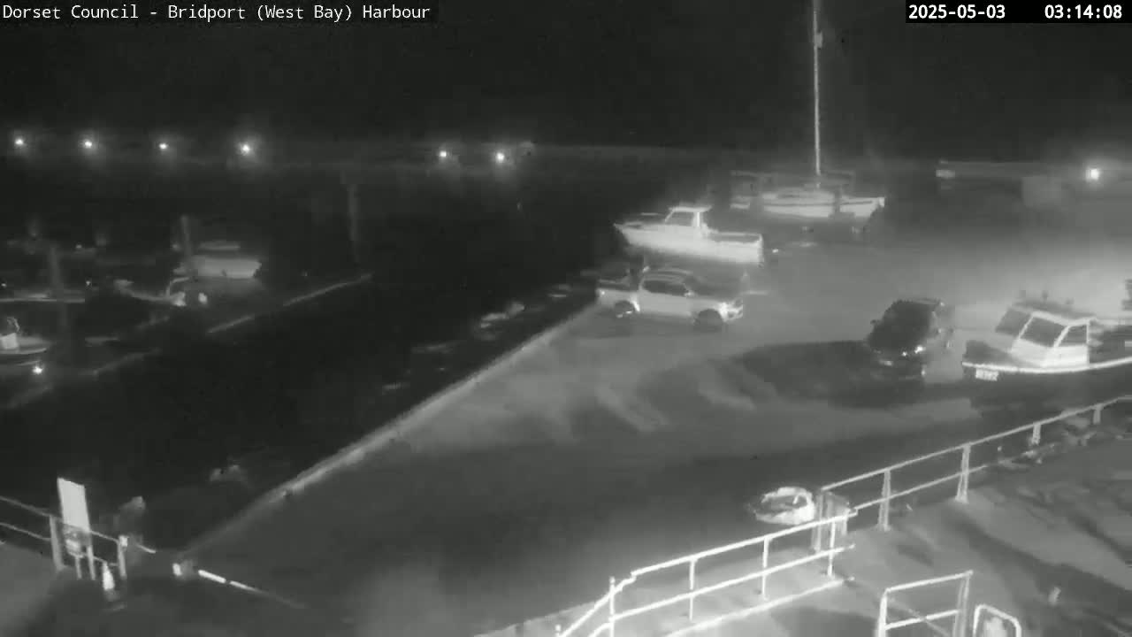 A nighttime, low-light harbor scene shows several boats and vehicles parked near a waterfront, with some boats on trailers.