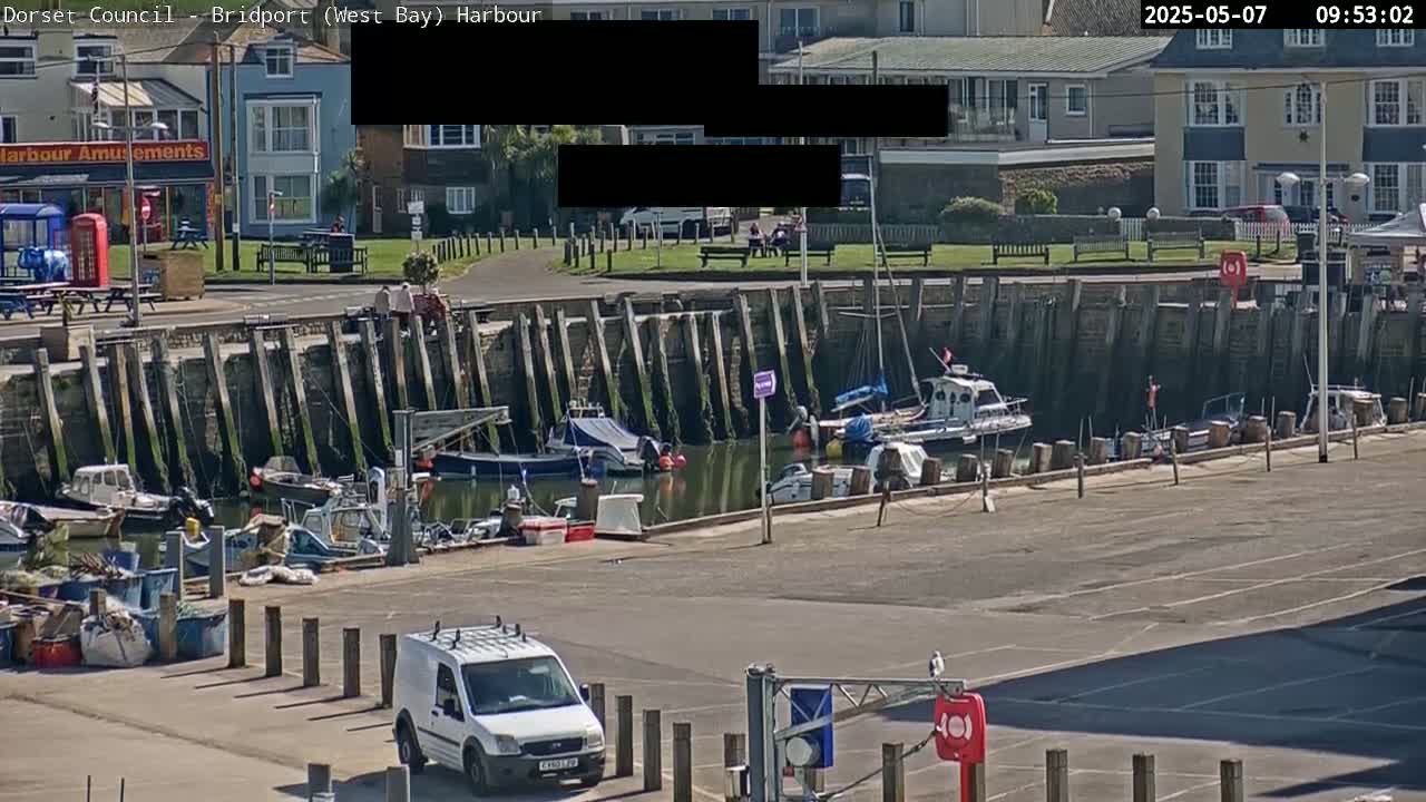 A harbor scene on a sunny day shows several boats docked along a seawall, with a white van parked in an empty lot and buildings visible in the background.