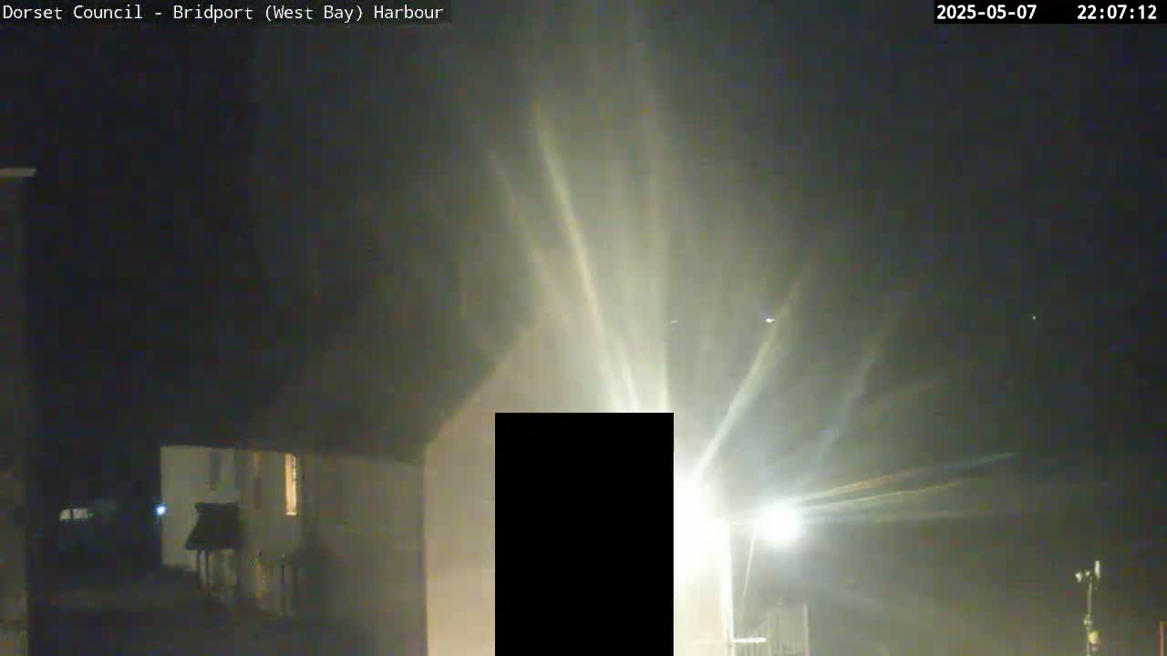 At night, bright light beams emanate from a central point, illuminating a partially visible building and a foggy or misty atmosphere.