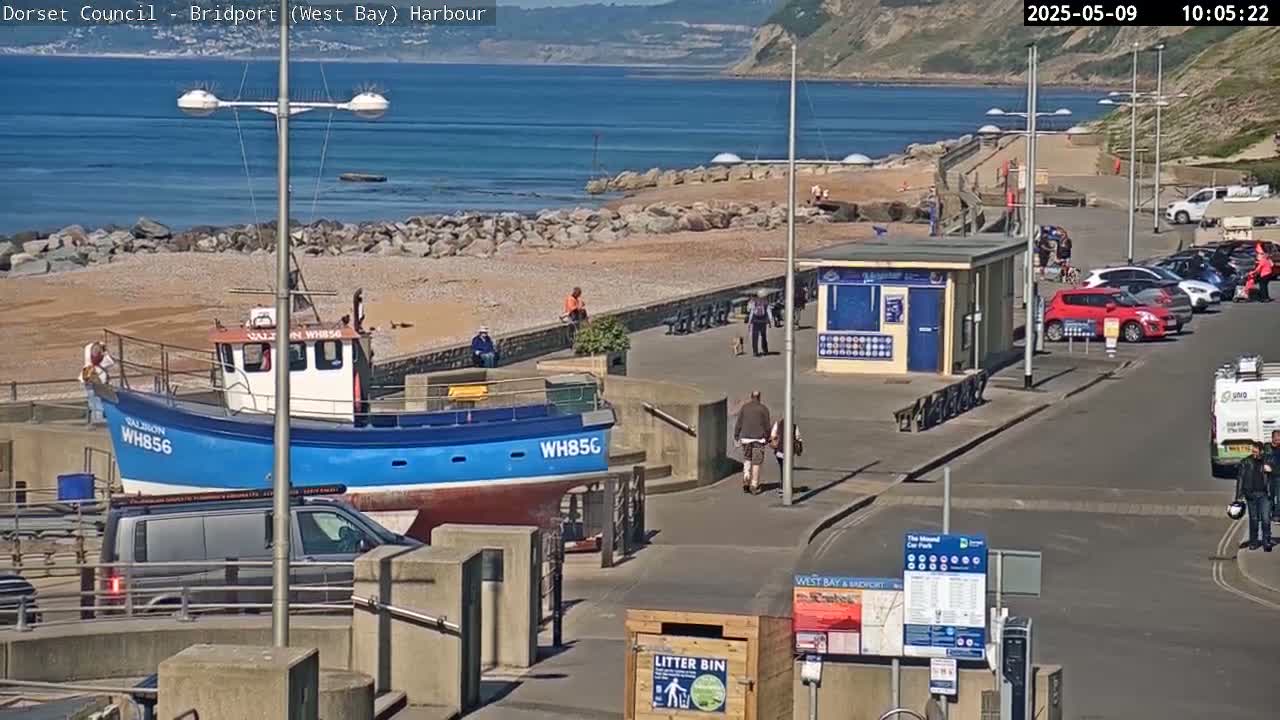A blue and white fishing boat sits in a harbor next to a pebble beach on a sunny day, with several people walking along the shoreline and cars parked nearby.