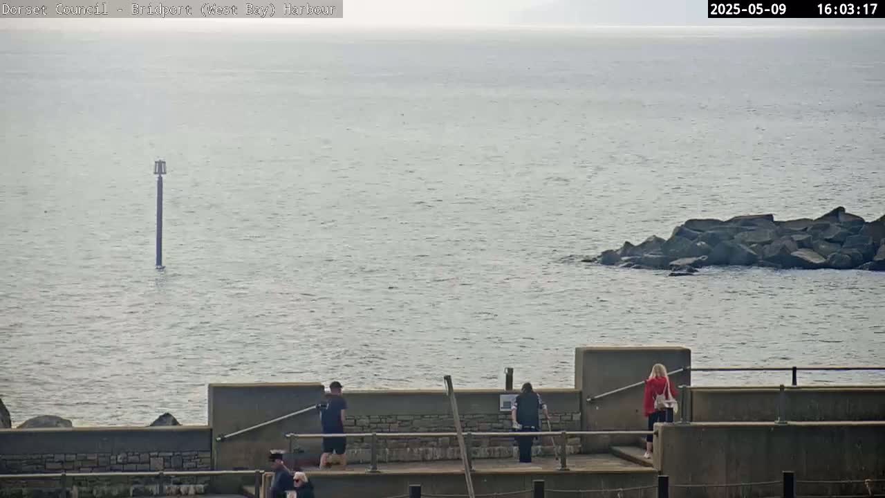 A calm body of water is seen from a high vantage point, with a few people standing on a low wall overlooking the water, a marker buoy in the distance, and a small rock formation jutting out of the water.
