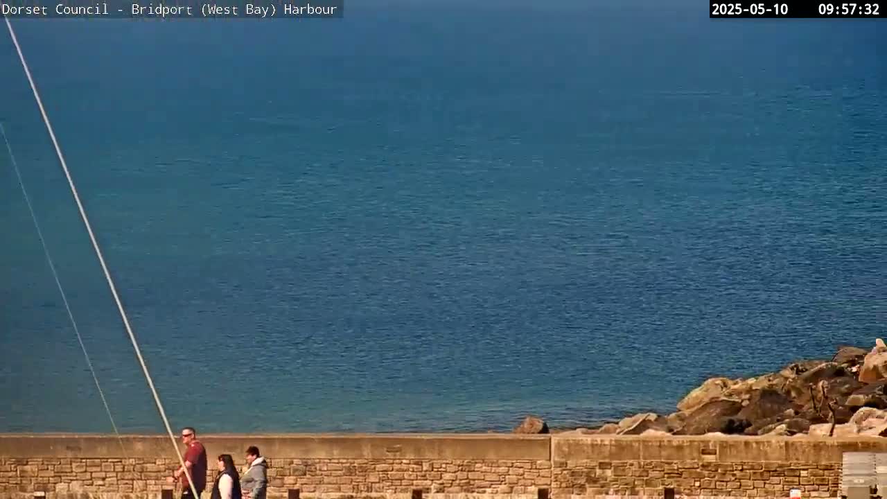 A calm, blue sea is visible beyond a low stone wall where three people are walking.