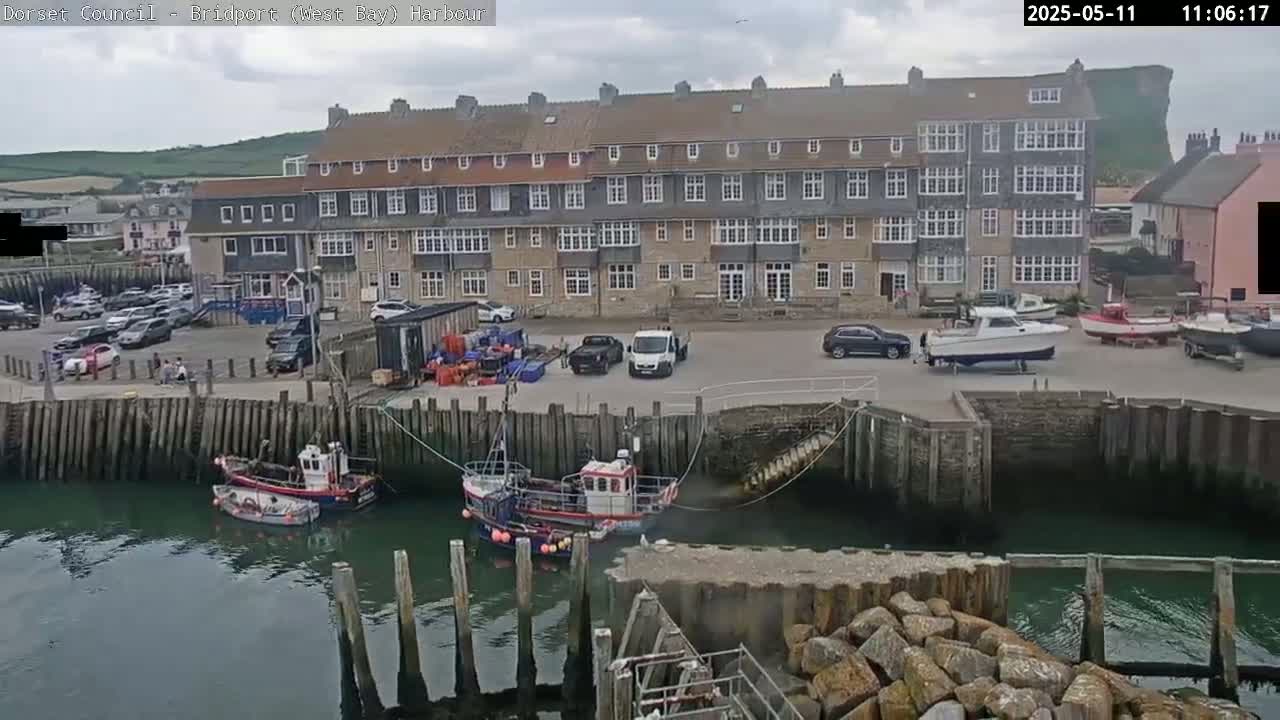 A multi-story building overlooks a harbor with several small boats and cars parked in a lot under partly cloudy skies.