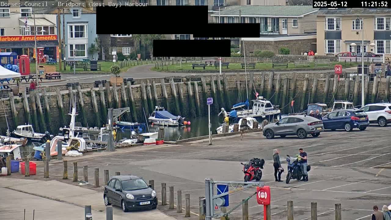 A harbor with several boats docked, cars parked in a lot, and people standing near motorcycles on an overcast day.