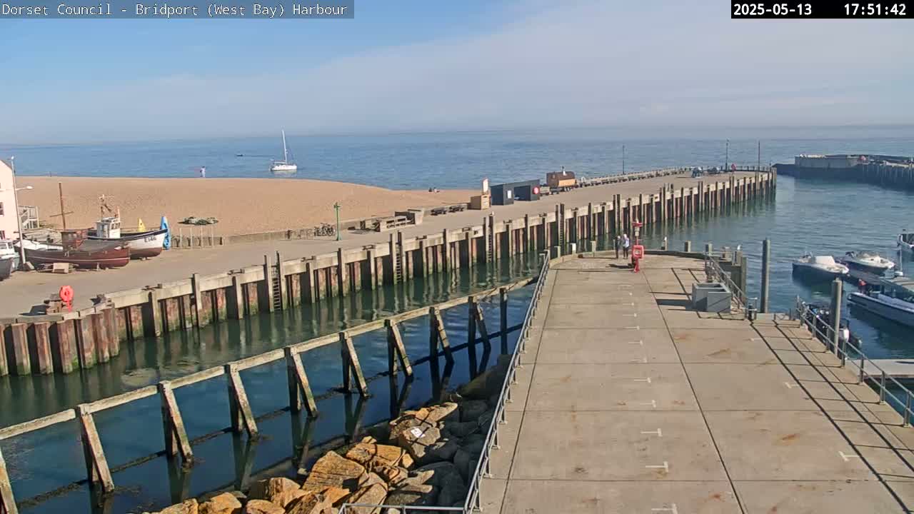 A harbor with a sandy beach, several boats, and a long concrete pier under a clear, sunny sky.