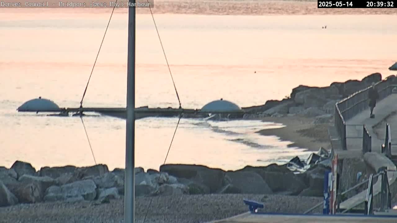 A calm, pink-hued sunset illuminates a rocky shoreline with two partially submerged, dome-shaped structures in the water near a pebble beach.